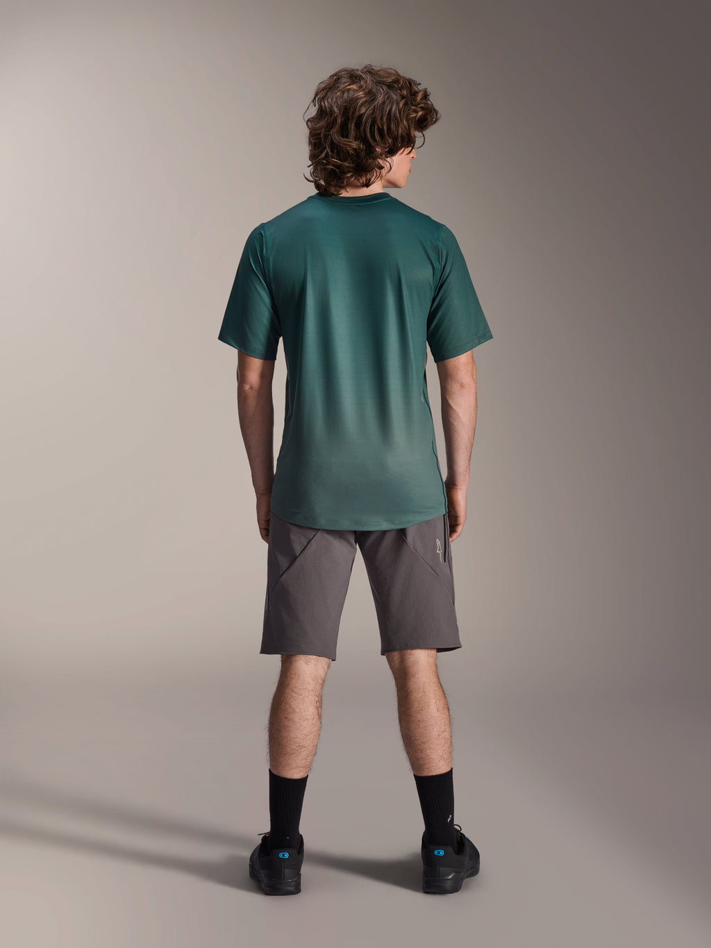 Alpinestars A-Dura Rise Jersey - Manga Corta, verde Silver Pine, vista posterior de un modelo masculino vistiendo la camiseta con pantalones cortos grises y calcetines negros, con una cola más larga y ajuste regular para ciclismo de montaña