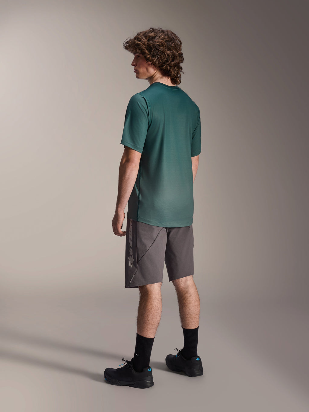 Alpinestars A-Dura Rise Jersey, jersey de MTB de manga corta, color verde Silver Pine, usado con pantalones cortos grises y zapatillas negras, vista trasera que muestra la cola más larga y el ajuste regular, tejido ligero que absorbe la humedad para ciclismo de montaña