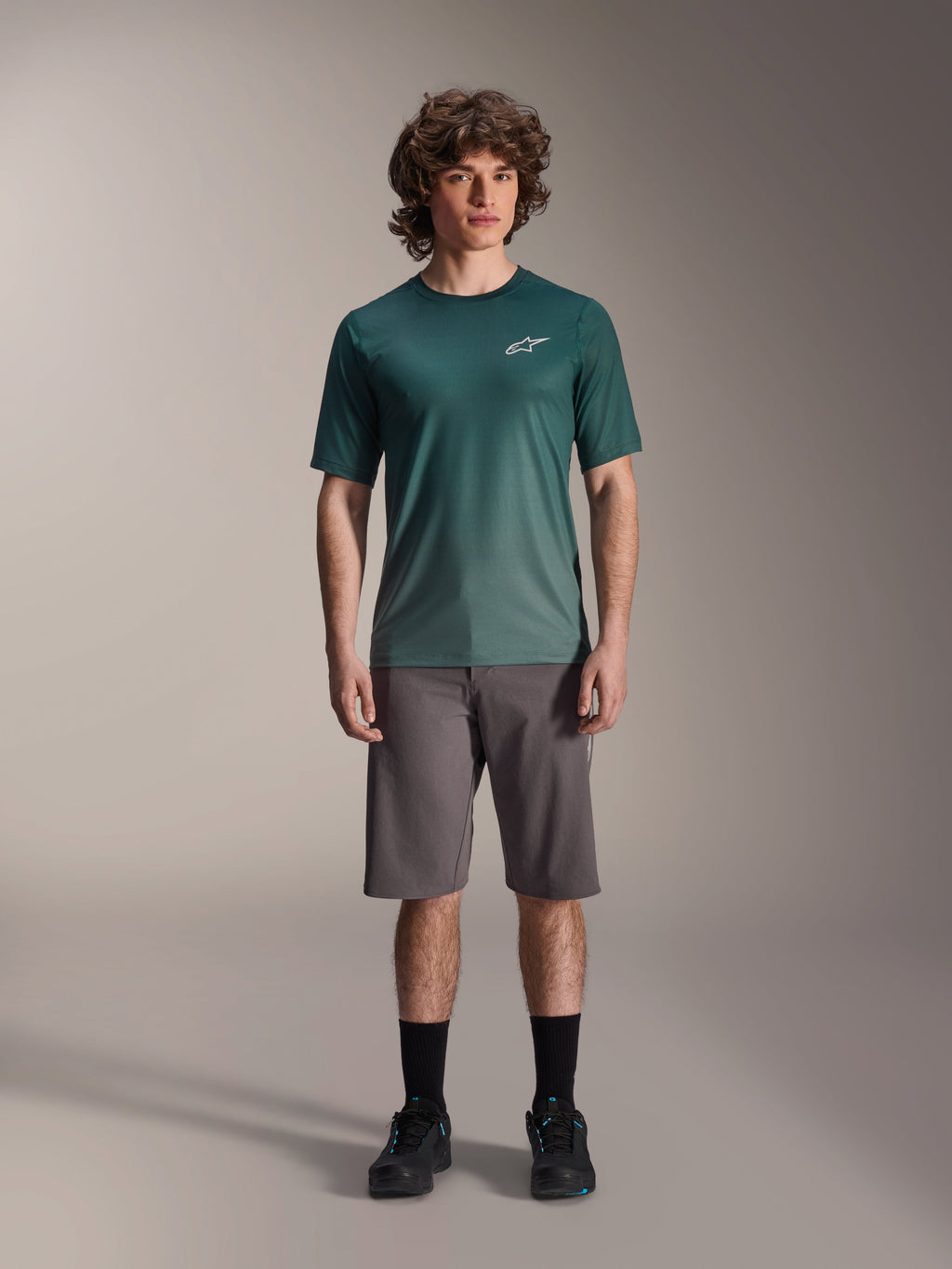 Modelo masculino con la camiseta Alpinestars A-Dura Rise de manga corta en Silver Pine, jersey de ciclismo verde con un pequeño logotipo blanco en el pecho, combinado con pantalones cortos grises de MTB y calzado negro, equipamiento casual para senderos