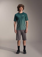 Modelo masculino con la camiseta Alpinestars A-Dura Rise de manga corta en Silver Pine, jersey de ciclismo verde con un pequeño logotipo blanco en el pecho, combinado con pantalones cortos grises de MTB y calzado negro, equipamiento casual para senderos