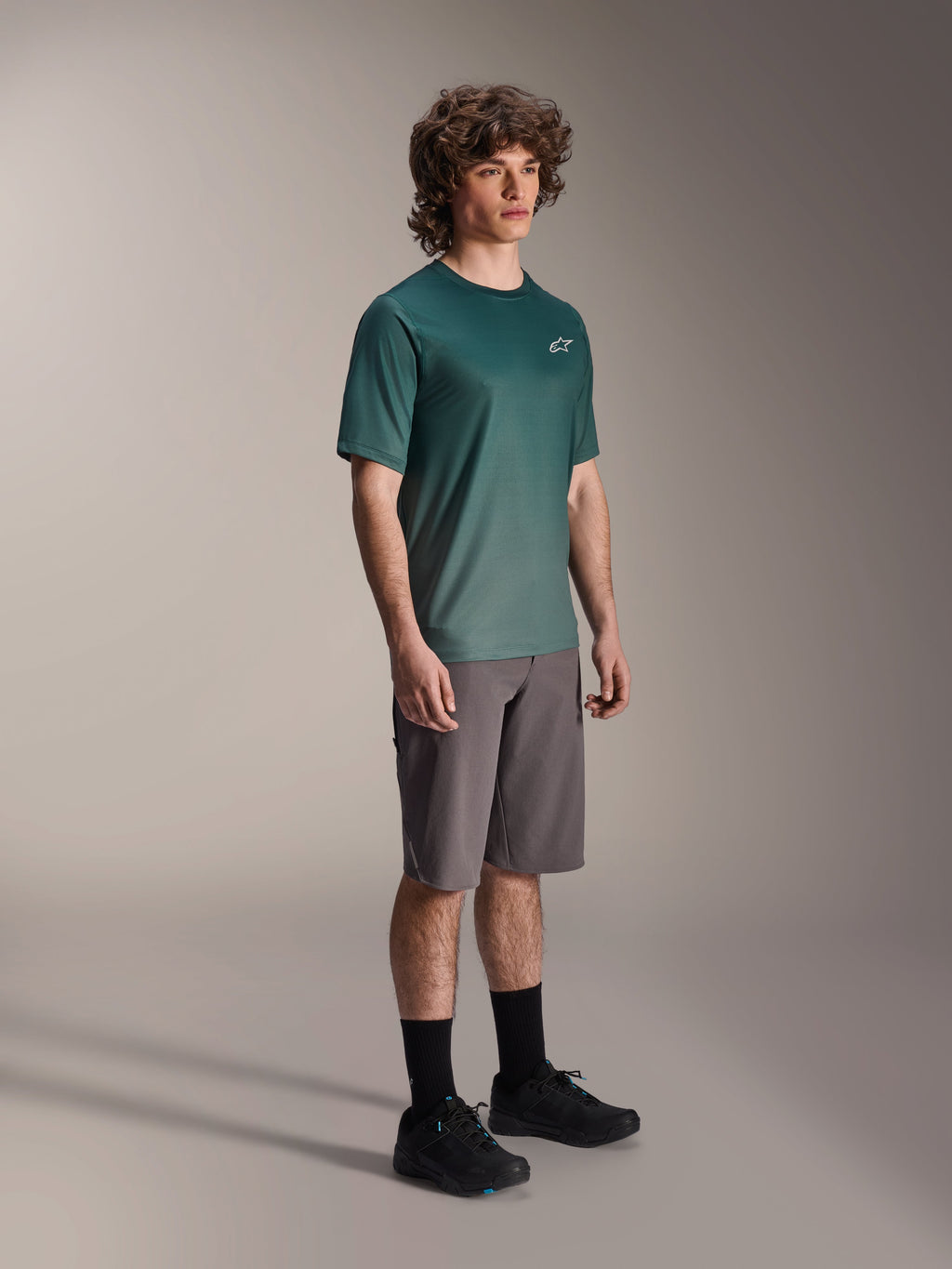 Alpinestars A-Dura Rise Jersey - Manga Corta en Silver Pine, jersey de ciclismo de montaña verde usado por un modelo con pantalones cortos grises y zapatillas negras, equipamiento casual para rutas de MTB con un pequeño logo blanco en el pecho