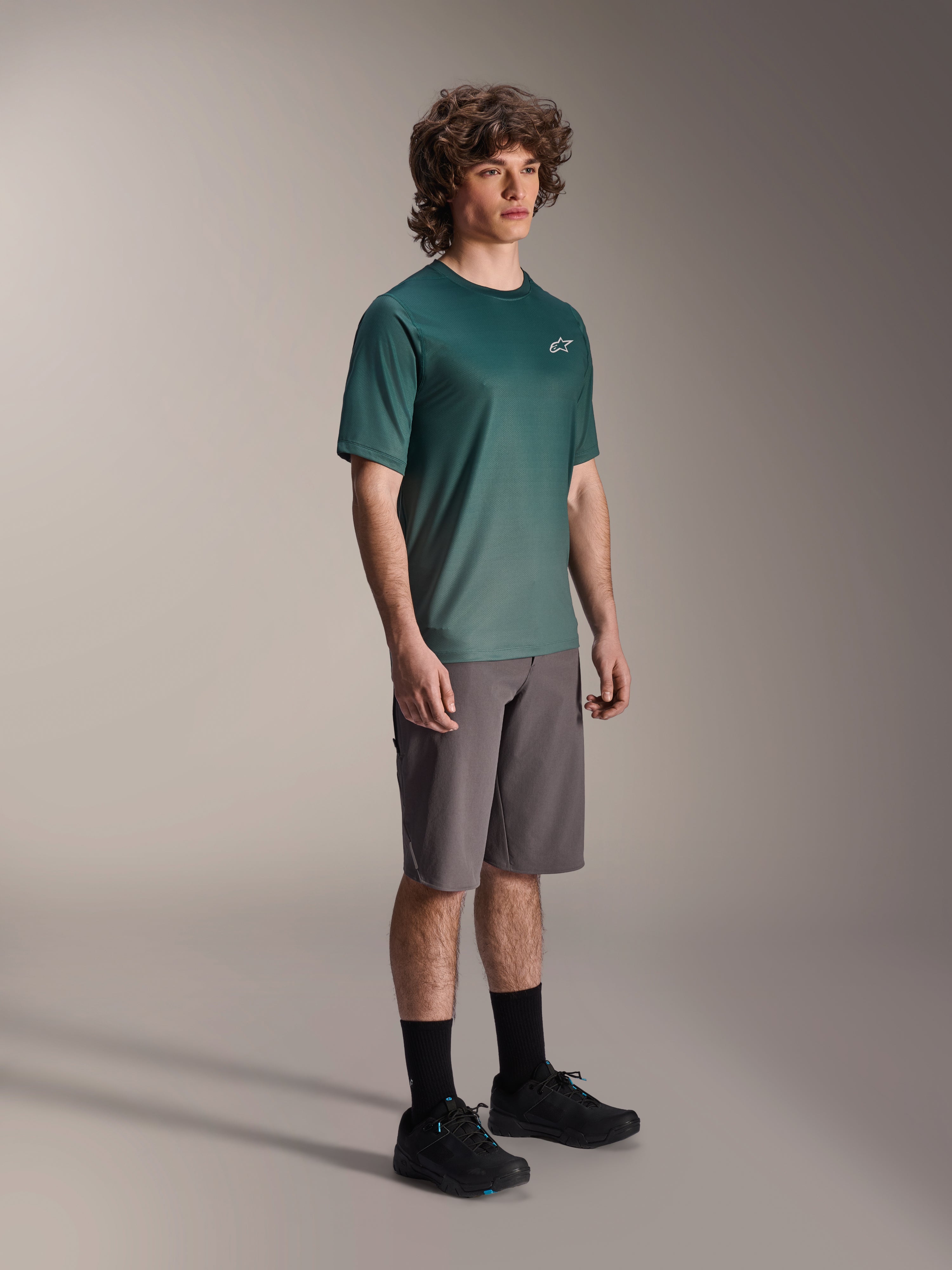 Alpinestars A-Dura Rise Jersey - Manga Corta en Silver Pine, jersey de ciclismo de montaña verde usado por un modelo con pantalones cortos grises y zapatillas negras, equipamiento casual para rutas de MTB con un pequeño logo blanco en el pecho