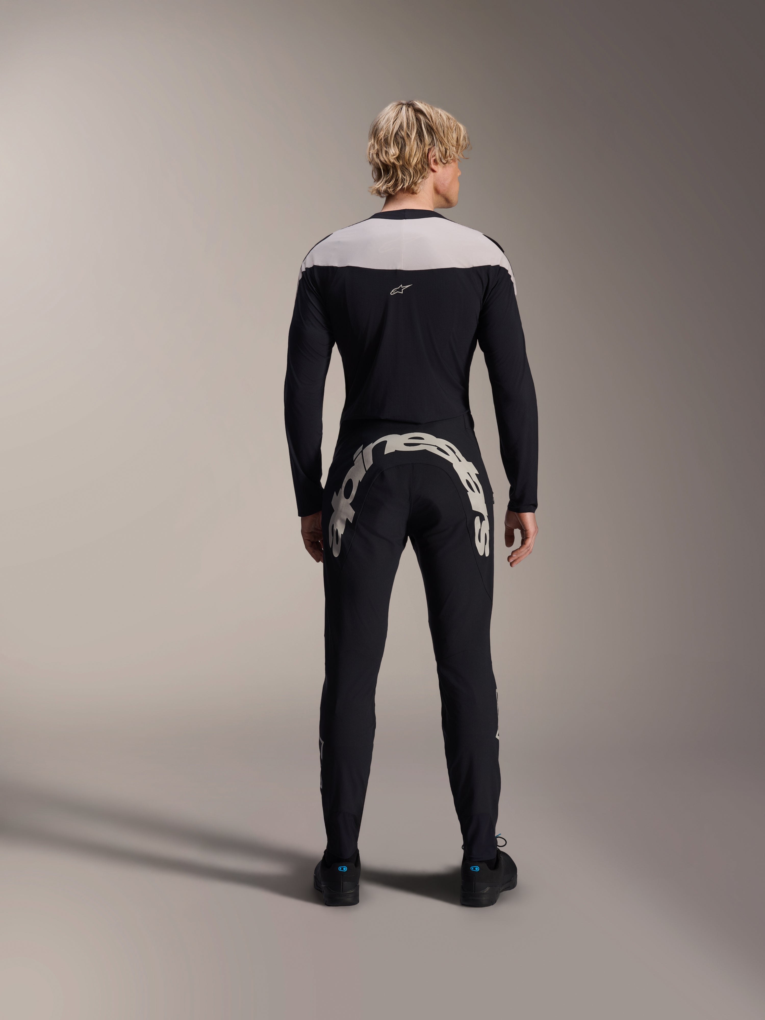 Hombre visto desde atrás con el Jersey Alpinestars A-Supra Melt de manga larga en negro, panel gris claro en los hombros, combinado con pantalones de MTB negros con un gran logotipo de Alpinestars en la parte trasera, kit de alto rendimiento para bicicleta de montaña