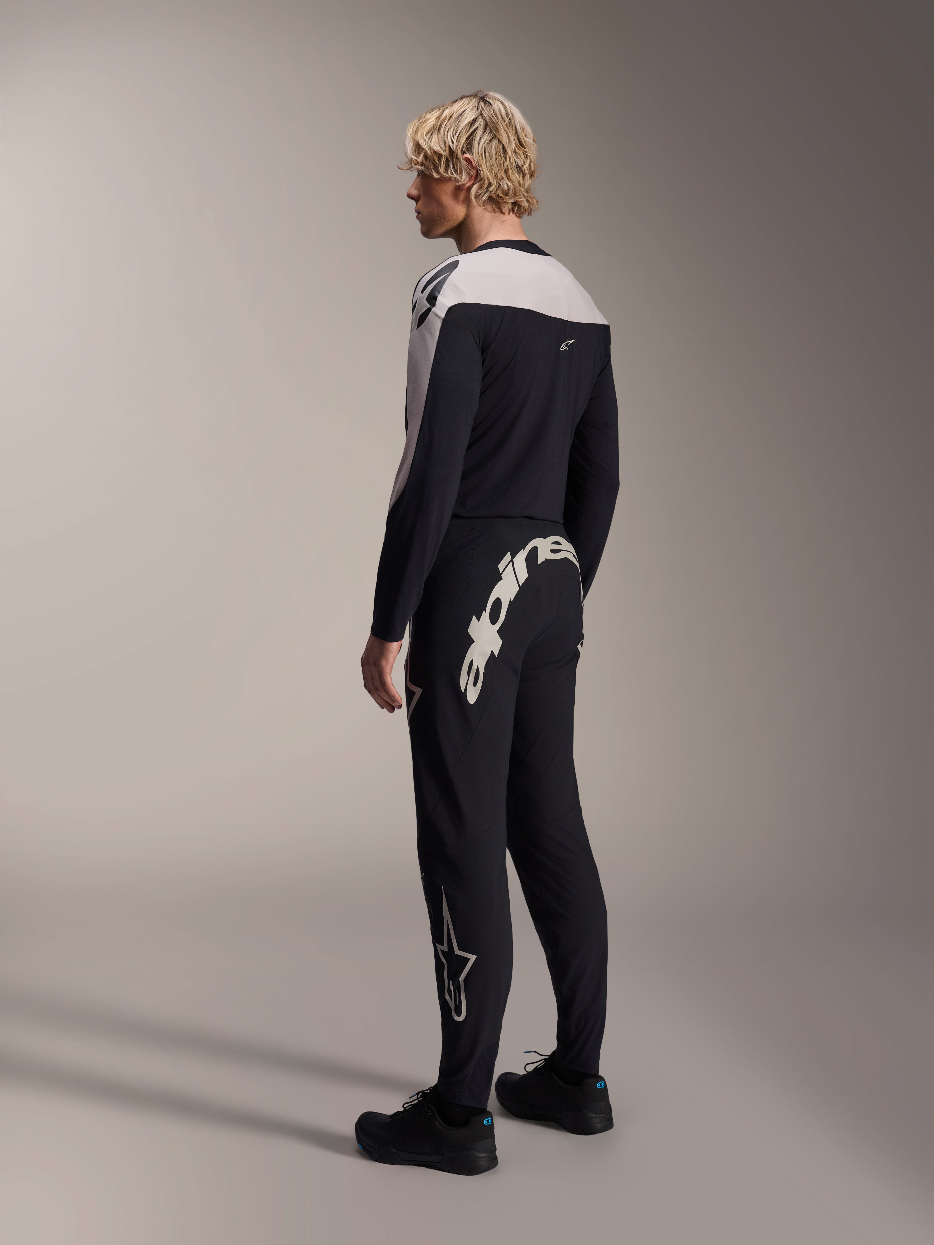 Pantalones Alpinestars A-Supra Melt, pantalones de MTB, negros, con una gran marca denominativa Alpinestars en blanco en el canesú trasero y el logotipo de la estrella en la pierna, usados por un modelo con un jersey de manga larga a juego y zapatillas negras, vista lateral trasera