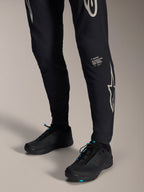 Alpinestars A-Supra Melt Pants, pantalones negros de mountain bike, con el logotipo y la marca denominativa de Alpinestars en blanco en la pierna, usados con zapatillas de ciclismo negras con detalles azules, equipamiento de alto rendimiento para todoterreno