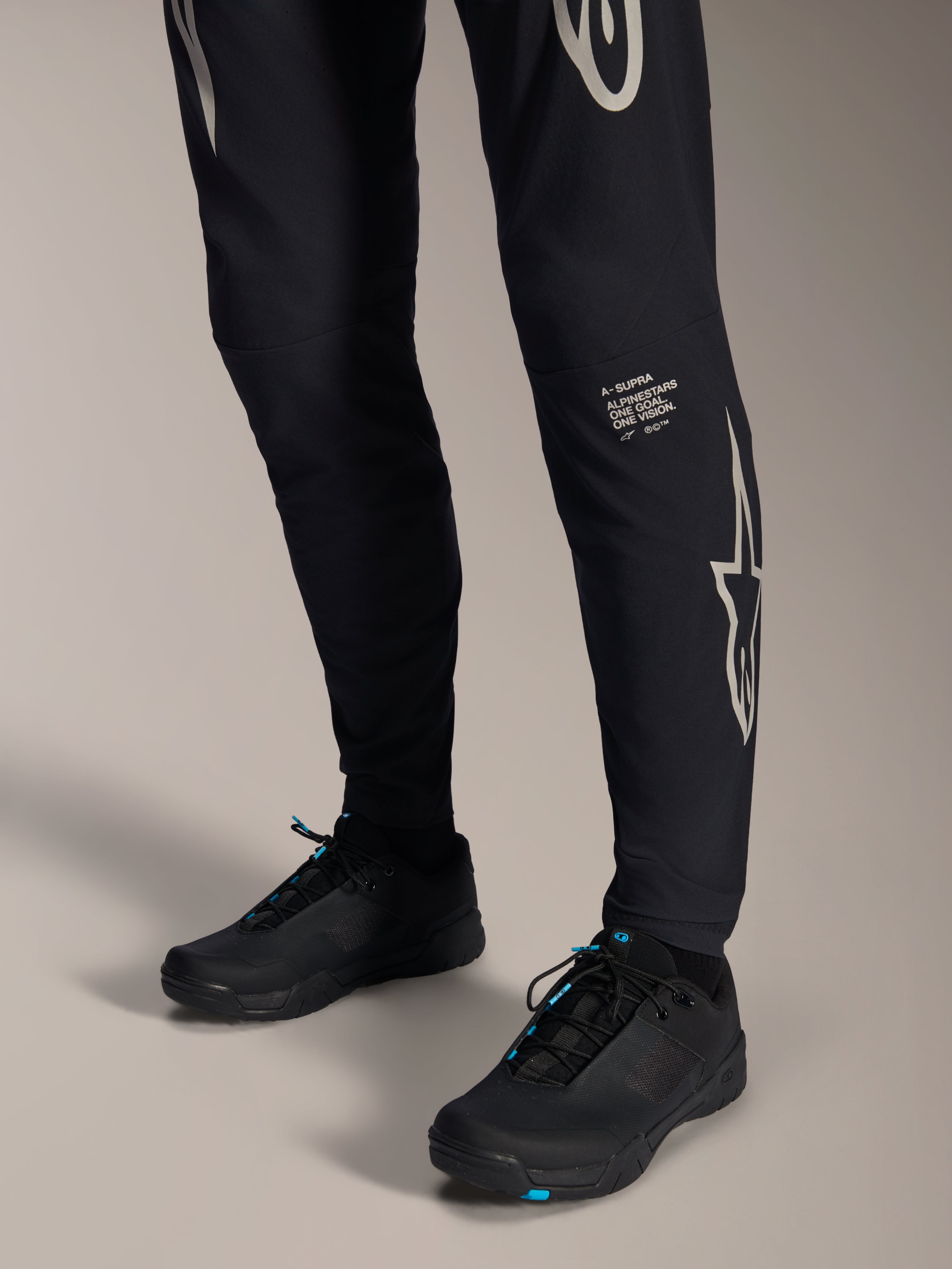 Alpinestars A-Supra Melt Pants, pantalones negros de mountain bike, con el logotipo y la marca denominativa de Alpinestars en blanco en la pierna, usados con zapatillas de ciclismo negras con detalles azules, equipamiento de alto rendimiento para todoterreno