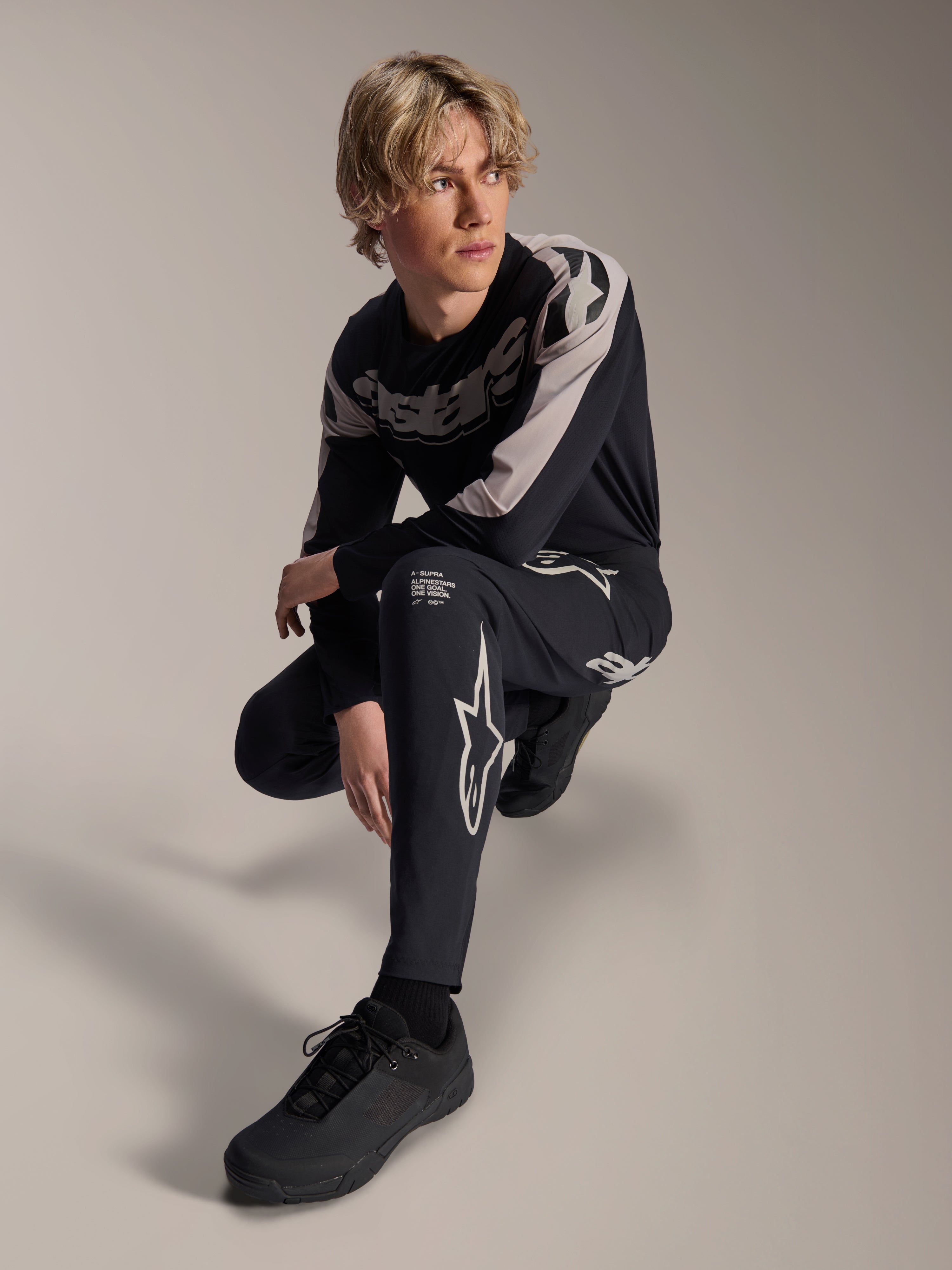 Pantalones y jersey de manga larga Alpinestars A-Supra Melt, negro y gris, con logotipos de estrella y marca destacados en blanco, lucidos por un modelo masculino en una pose agachada, equipamiento coordinado de carreras MTB