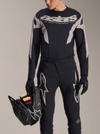 Kit de carreras MTB Alpinestars A-Supra Melt, jersey de manga larga y pantalones, negro y gris con logotipos Astars blancos, modelo sosteniendo un casco integral negro y usando guantes a juego, equipamiento de alto rendimiento para ciclismo de montaña, descenso y enduro