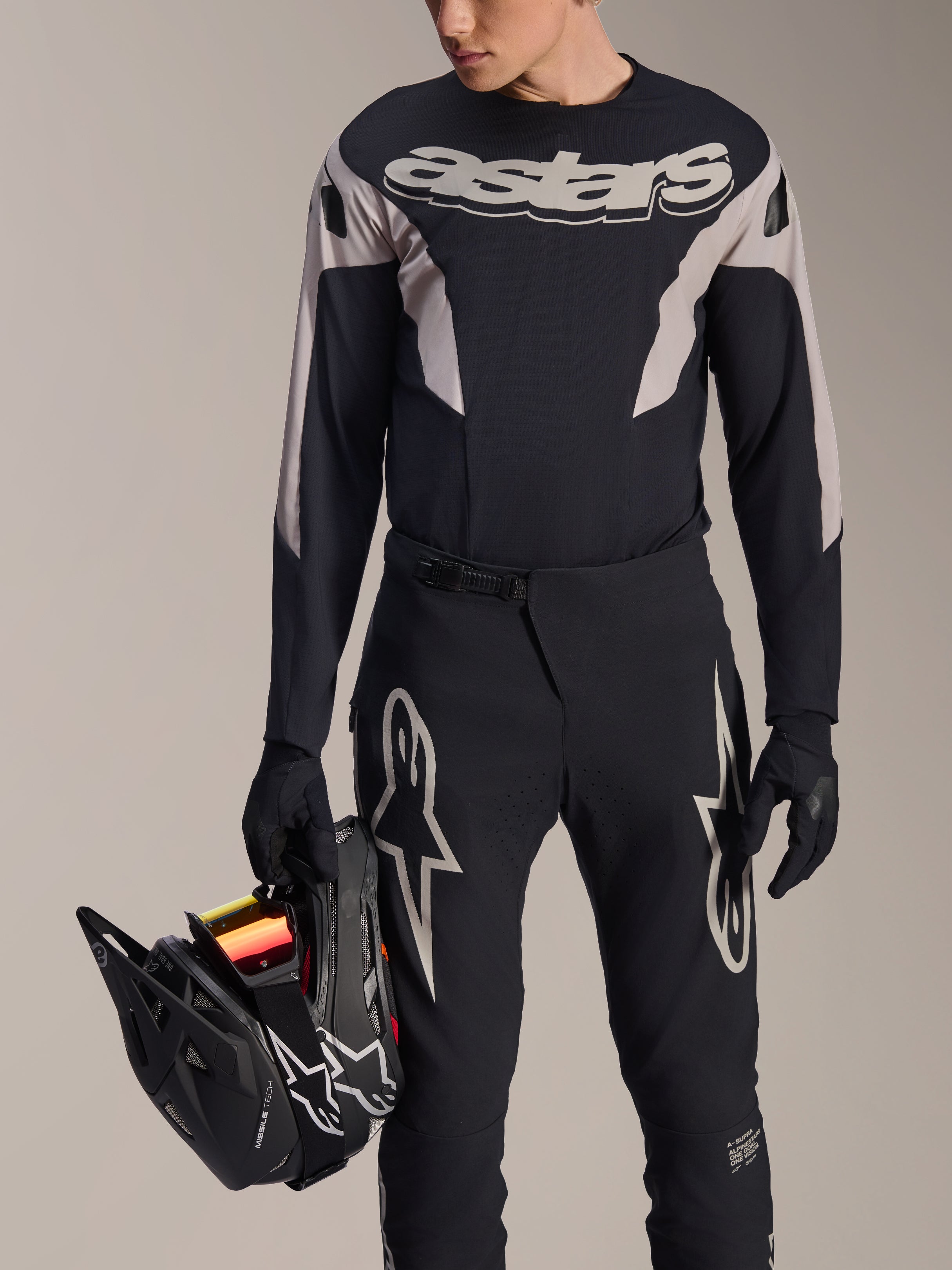 Jersey Alpinestars A-Supra Melt de manga larga, negro con paneles gráficos grises y logotipo 'astars' en el pecho, usado con pantalones y guantes de MTB negros mientras sostiene un casco integral negro, equipo de competición de MTB de alto rendimiento