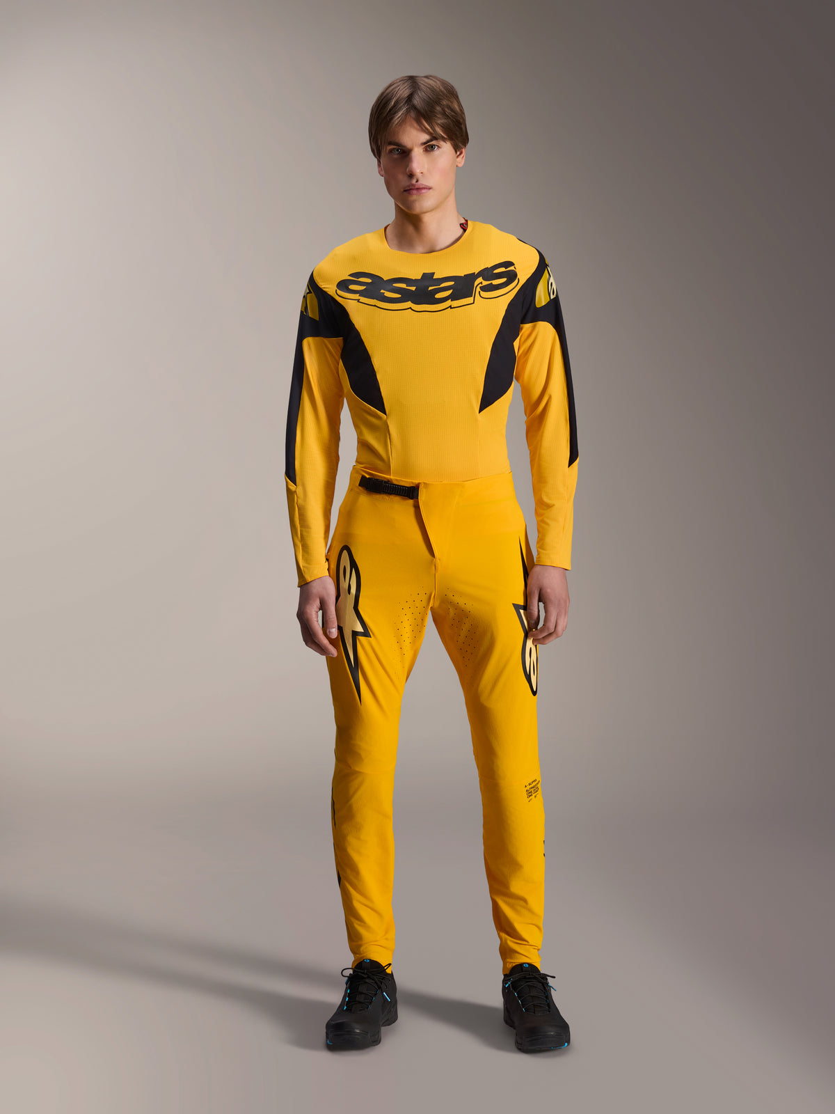 Limited Edition A-Supra Melt Gold Gear Set, Alpinestars Mountain Bike Apparel, Dark Yellow and Black, Model wearing long-sleeve jersey with bold 'astars' chest logo and matching performance pants, featuring laser-cut ventilation on inner thighs and a secure ratchet waist closure, professional MTB racing kit
