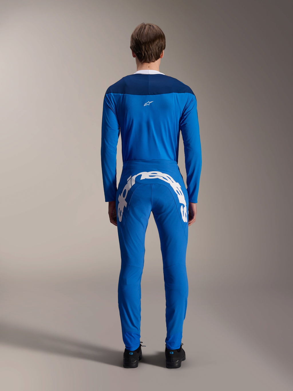 Pantalones Alpinestars A-Supra Melt, azul, vista trasera con un gran logotipo de Alpinestars en blanco en el canesú trasero, combinados con un jersey de manga larga azul a juego y calzado negro, equipamiento de competición de MTB de alto rendimiento