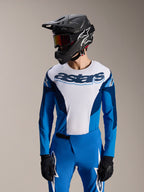 Modelo con el Jersey Alpinestars A-Supra Melt de manga larga en azul y blanco, con el logotipo 'astars' en el pecho, combinado con pantalones de MTB azules a juego, casco integral negro y gafas, conjunto completo de equipamiento para ciclismo de montaña.