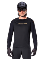 Jersey de manga larga Alpinestars A-Aria Polartec Switch, negro, logotipo de Alpinestars en dorado en el pecho, usado con un casco de MTB negro, gafas de sol oscuras y guantes negros, equipamiento profesional para trail en bicicleta de montaña