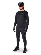 Jersey Alpinestars A-Aria Polartec Switch de manga larga, negro, logotipo en bronce en el pecho, usado por un ciclista de montaña con casco negro y gafas de sol, ropa de ciclismo de alto rendimiento para trail