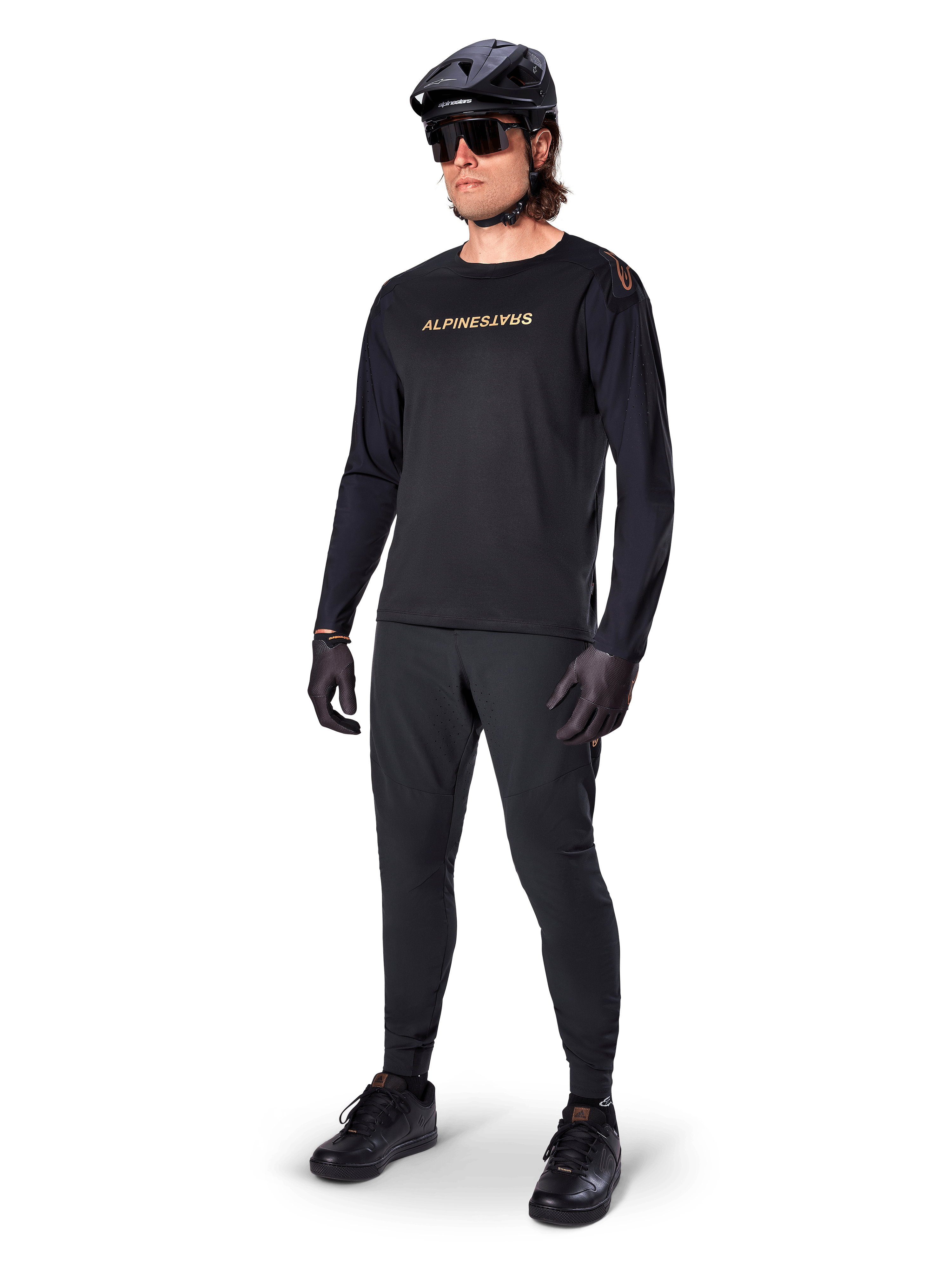 Jersey Alpinestars A-Aria Polartec Switch de manga larga, negro, logotipo en bronce en el pecho, usado por un ciclista de montaña con casco negro y gafas de sol, ropa de ciclismo de alto rendimiento para trail