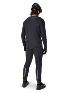 Vista trasera de un ciclista de montaña con el jersey de manga larga Alpinestars A-Aria Polartec Switch en negro, combinado con pantalones de MTB negros con la marca Alpinestars en color tostado en las pantorrillas, casco y guantes negros, equipamiento de ciclismo profesional para senderos todoterreno.