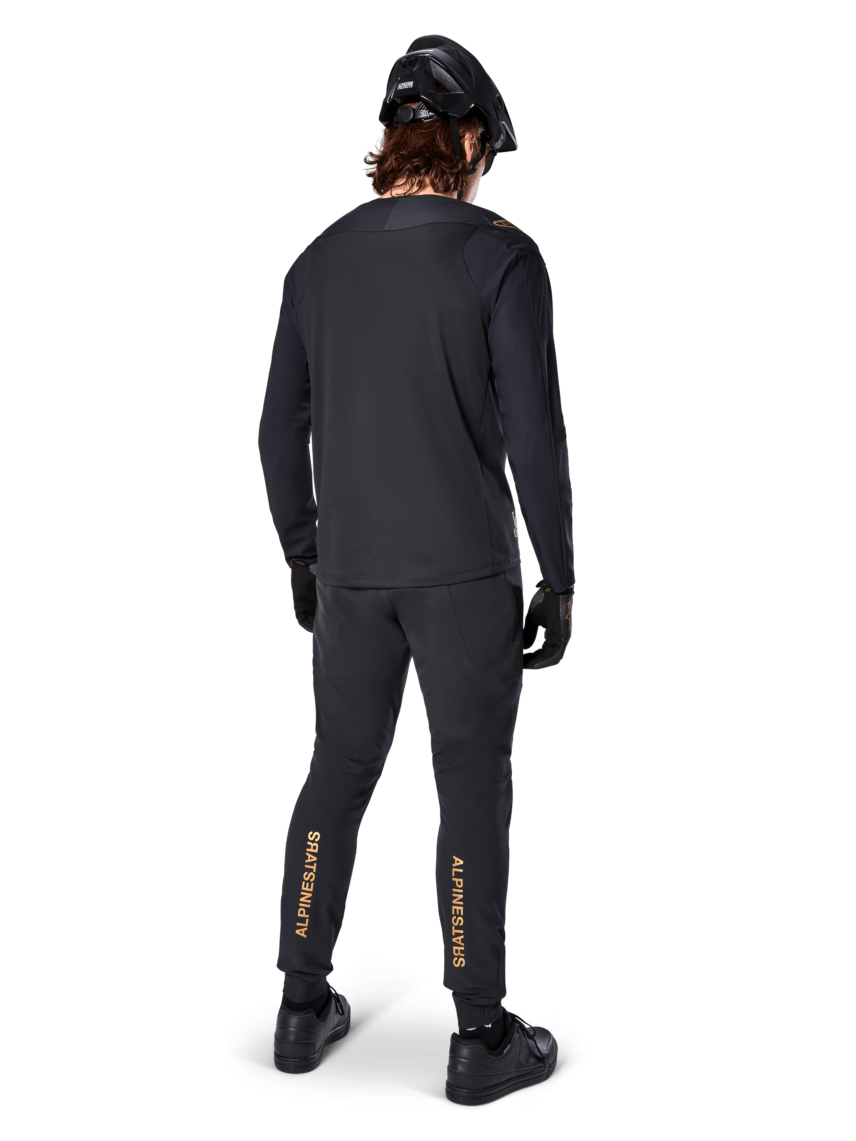 Vista trasera de un ciclista de montaña con el jersey de manga larga Alpinestars A-Aria Polartec Switch en negro, combinado con pantalones de MTB negros con la marca Alpinestars en color tostado en las pantorrillas, casco y guantes negros, equipamiento de ciclismo profesional para senderos todoterreno.