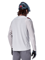 Jersey de manga larga Alpinestars A-Aria Polartec Switch, gris claro, visto desde atrás en un ciclista, usado con un casco de montaña azul y pantalones grises, ropa de rendimiento para ciclismo de montaña en senderos todoterreno