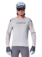 Alpinestars A-Aria Polartec Switch Jersey de manga larga, jersey de ciclismo de montaña en color gris claro con perforaciones láser en las mangas y logotipo en el pecho, usado con casco azul claro, gafas de sol y guantes grises