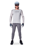 Jersey Alpinestars A-Aria Polartec Switch de manga larga, gris claro, usado por un ciclista de montaña con pantalones de MTB gris oscuro, casco azul claro y gafas de sol, con logotipo en el pecho y ventilación perforada por láser en las mangas, equipamiento completo para ciclismo todoterreno