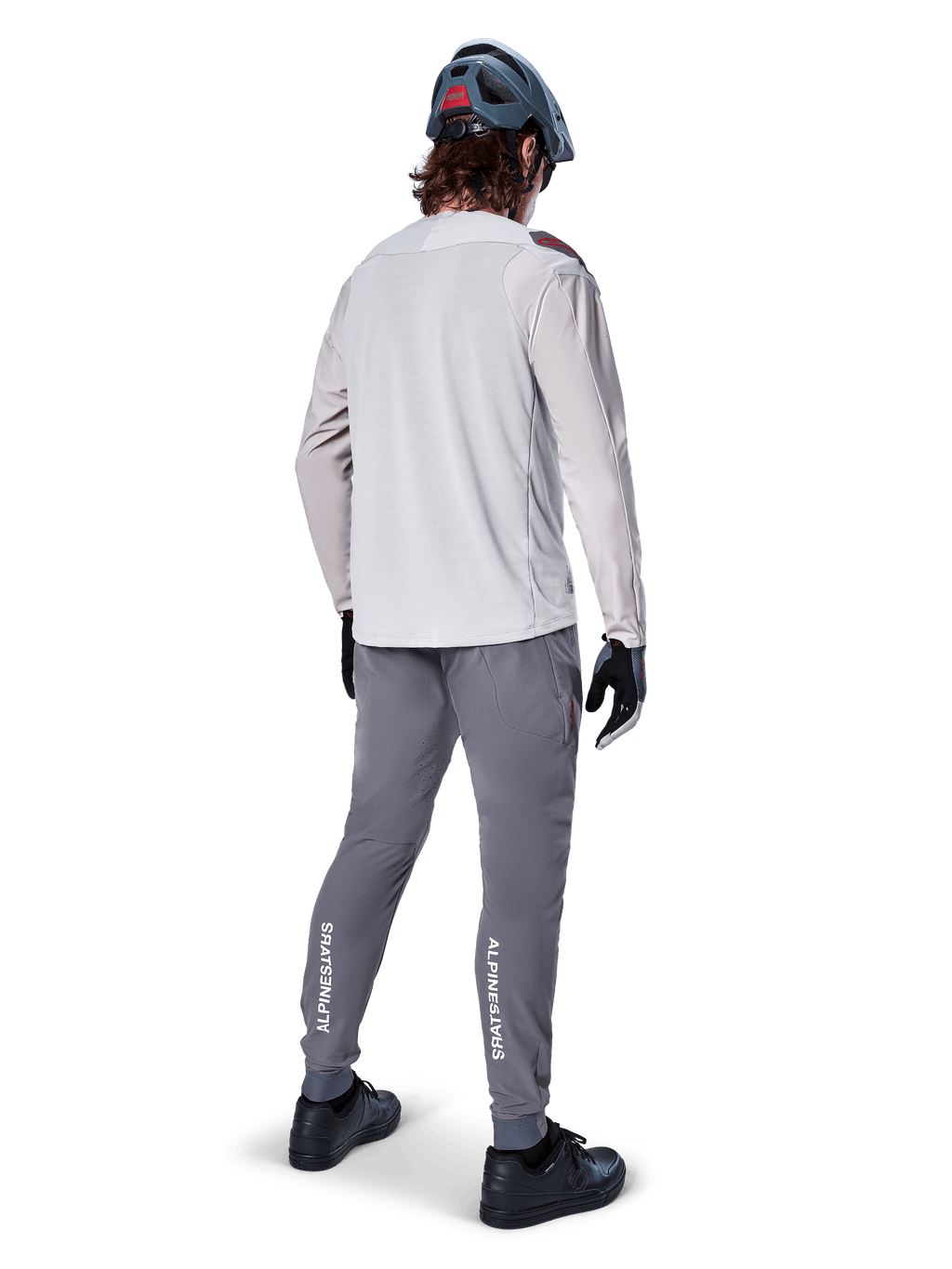 Vista trasera de un ciclista con el jersey de manga larga Alpinestars A-Aria Polartec Switch en gris claro, combinado con pantalones de montaña gris oscuro con la marca Alpinestars en blanco en las pantorrillas, guantes negros y casco gris.
