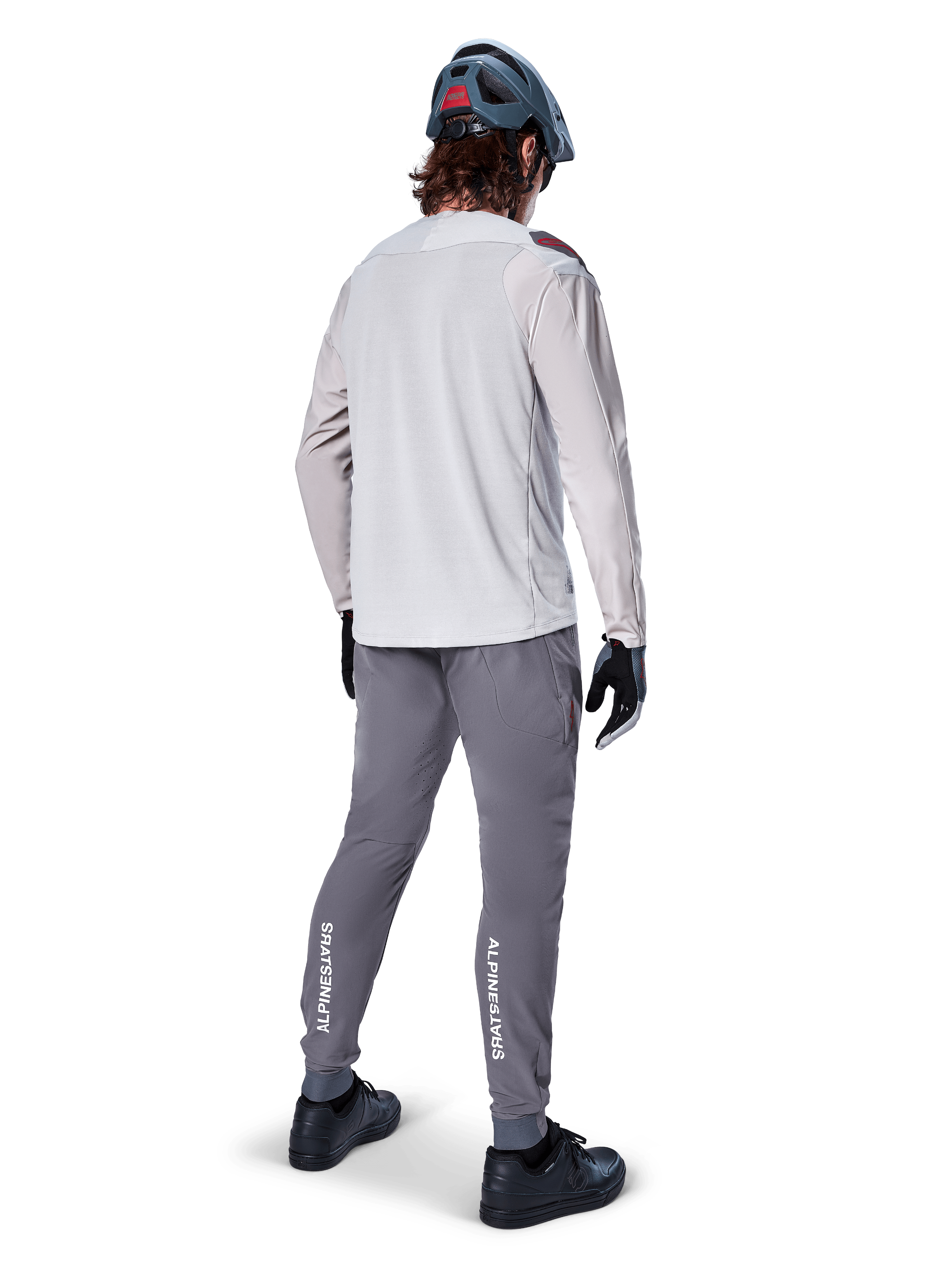 Vista trasera de un ciclista con el jersey de manga larga Alpinestars A-Aria Polartec Switch en gris claro, combinado con pantalones de montaña gris oscuro con la marca Alpinestars en blanco en las pantorrillas, guantes negros y casco gris.