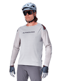 Alpinestars A-Aria Polartec Switch Jersey de manga larga, camiseta de ciclismo gris claro con logotipo negro en el pecho y detalles rojos en los hombros, mangas perforadas con láser para ventilación, combinada con casco azul claro, gafas de sol y pantalones grises, ropa técnica de montaña para trail
