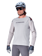 Alpinestars A-Aria Polartec Switch Jersey de manga larga, camiseta de ciclismo gris claro con logotipo negro en el pecho y detalles rojos en los hombros, mangas perforadas con láser para ventilación, combinada con casco azul claro, gafas de sol y pantalones grises, ropa técnica de montaña para trail
