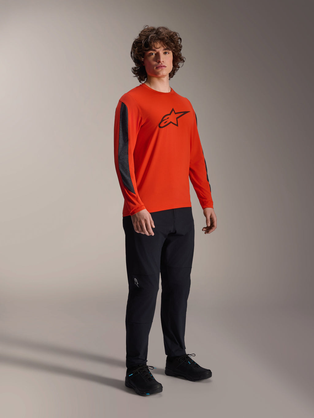 Modelo masculino vistiendo el jersey Alpinestars A-Dura Insert Astar de manga larga, color naranja Spicy Orange con logotipo Astar negro en el pecho e inserciones gris oscuro en las mangas, usado con pantalones negros, equipamiento profesional de ciclismo MTB