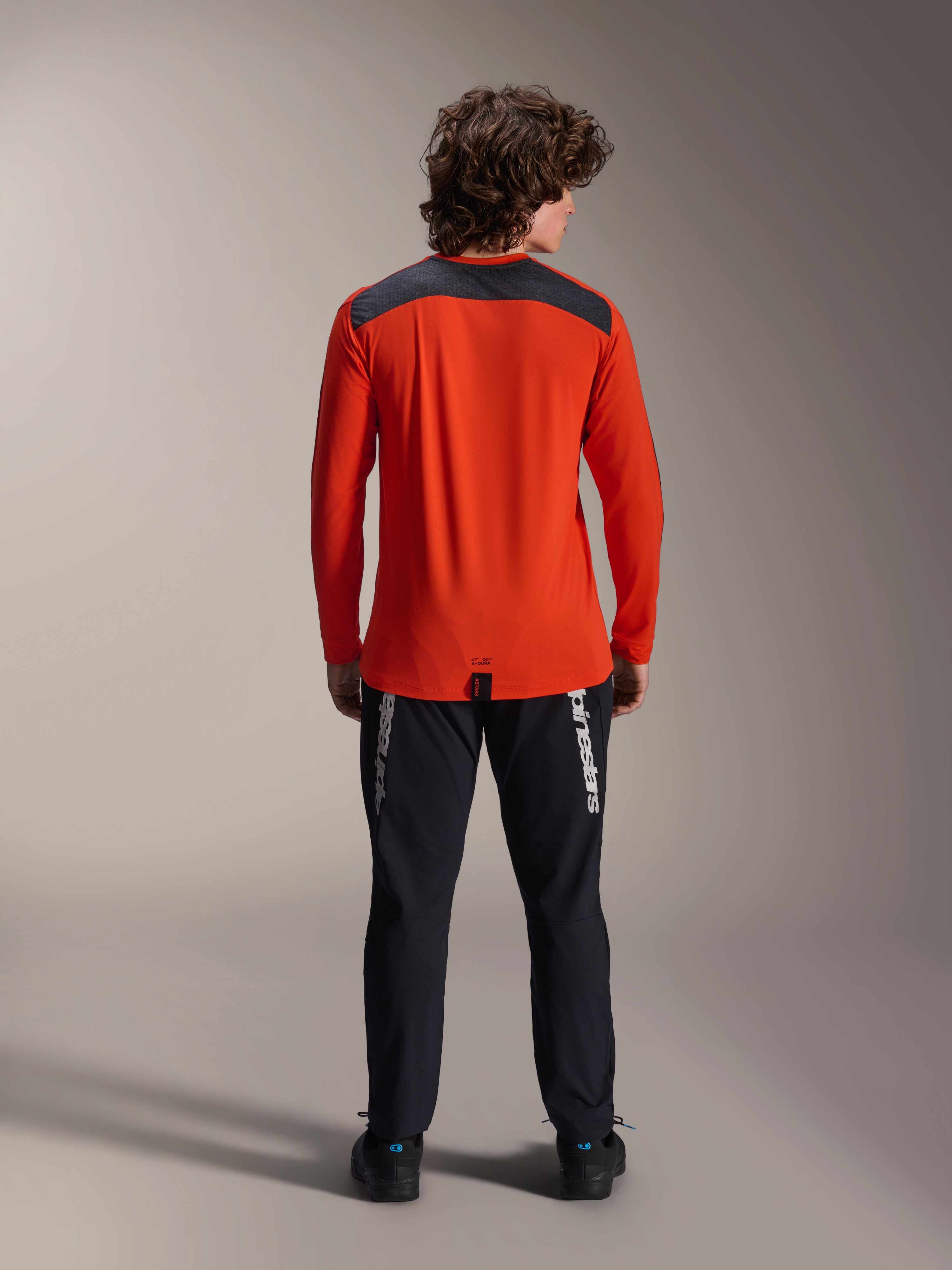 Jersey Alpinestars A-Dura Insert Astar de manga larga en Spicy Orange, vista trasera que muestra el panel de los hombros en gris carbón, usado con pantalones Alpinestars negros y calzado negro, fotografía de estudio profesional de equipamiento de alto rendimiento para MTB