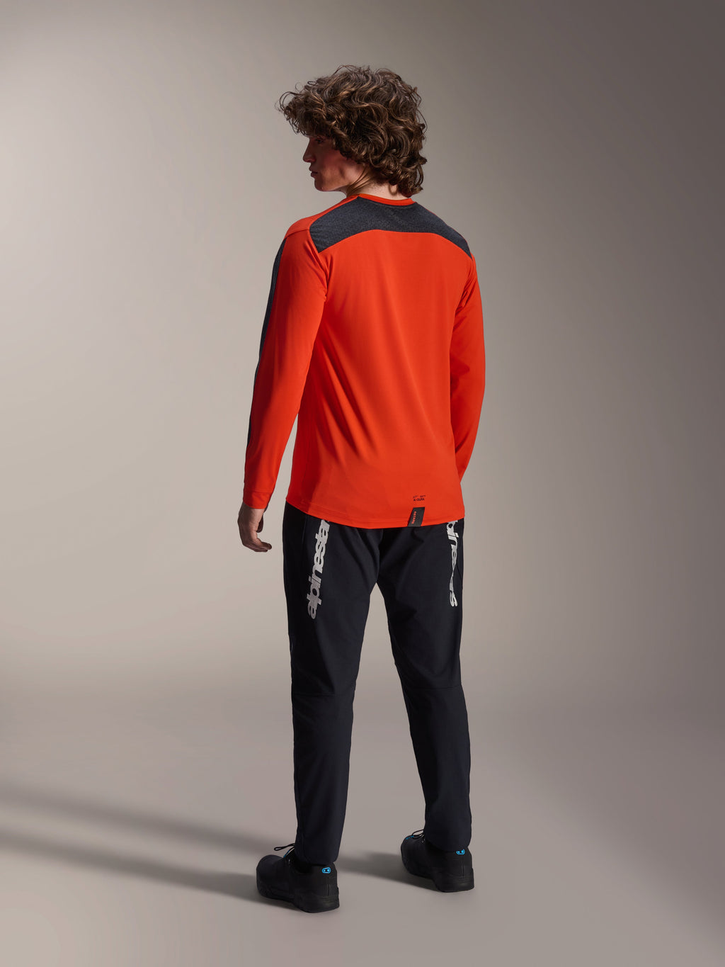 Vista posterior de un modelo vistiendo el jersey Alpinestars A-Dura Insert Astar en Spicy Orange, jersey de MTB de manga larga con panel gris oscuro en los hombros, combinado con pantalones negros con logo de la marca, equipamiento de ciclismo todoterreno.