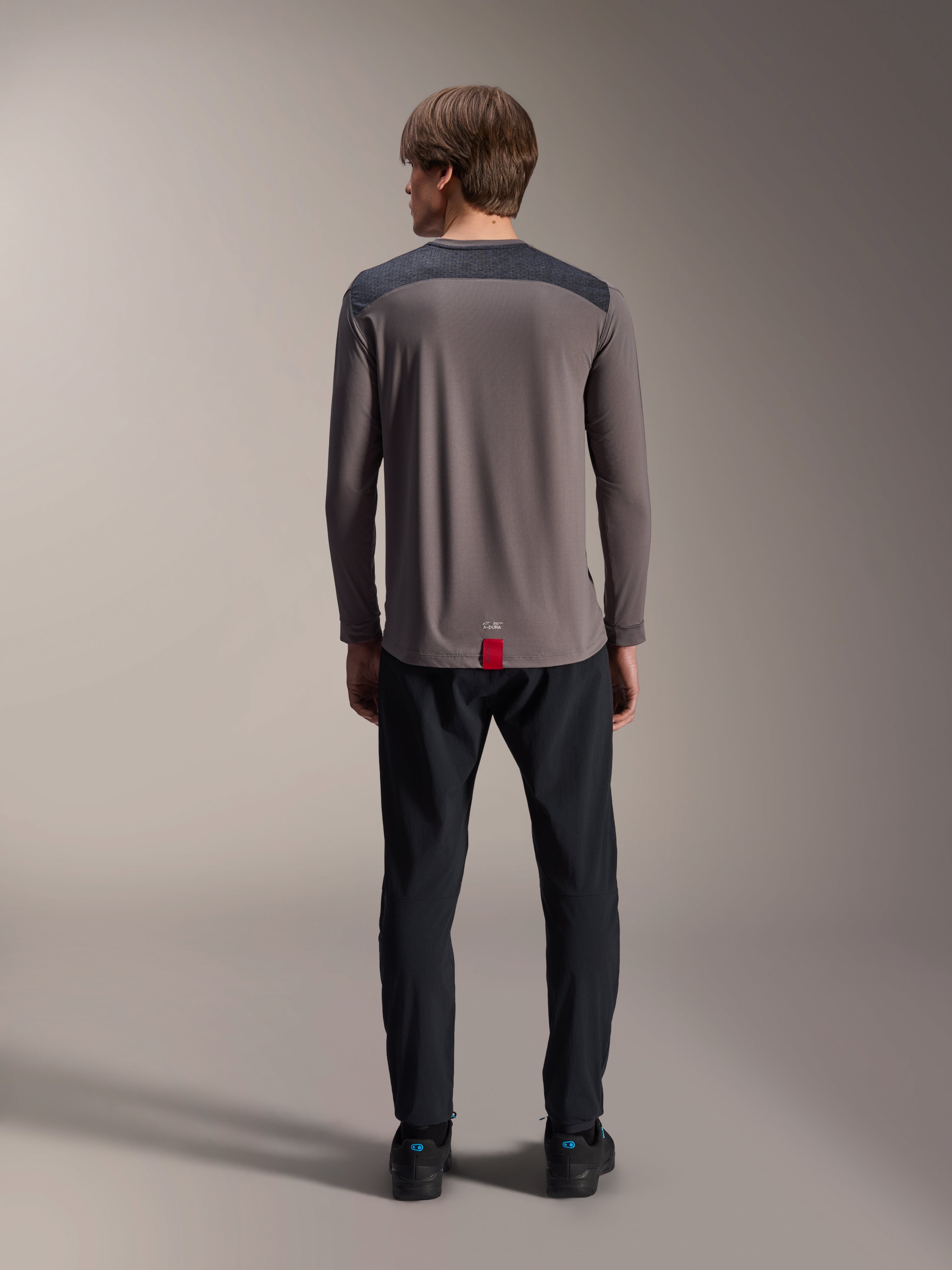 Alpinestars A-Dura Insert Astar Long Sleeve Jersey en gris oscuro, vista trasera, con paneles de color carbón en los hombros y una etiqueta roja en el dobladillo, combinado con pantalones y calzado negros, ropa técnica para bicicleta de montaña