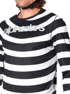 Primer plano del jersey de manga larga Alpinestars A-Aria Oscar en color negro, con rayas blancas horizontales, diseño gráfico en espiral de Oscar y logotipo plateado reflectante en el pecho, vestido por un ciclista con la correa del casco visible.