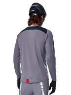 Alpinestars A-Dura Dri Astar Long Sleeve Jersey, jersey de MTB gris oscuro visto desde atrás, con un panel negro en los hombros y una pequeña etiqueta roja de la marca, usado con un casco de bicicleta de montaña y guantes negros.