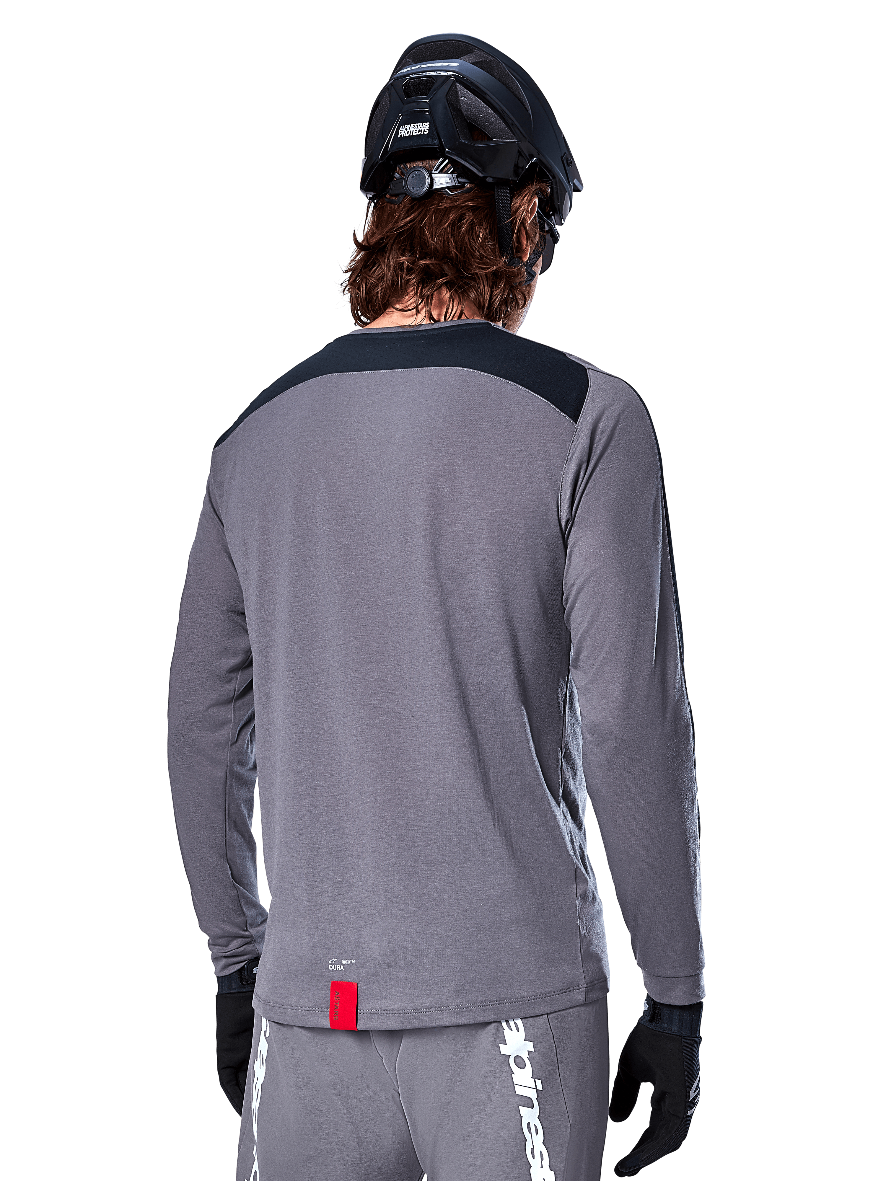 Alpinestars A-Dura Dri Astar Long Sleeve Jersey, jersey de MTB gris oscuro visto desde atrás, con un panel negro en los hombros y una pequeña etiqueta roja de la marca, usado con un casco de bicicleta de montaña y guantes negros.