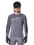 Jersey de manga larga Alpinestars A-Dura Dri Astar, gris oscuro, con un gran logotipo Astar blanco en el pecho y paneles de malla negros en los brazos, usado por un ciclista con casco negro de bicicleta de montaña y gafas de sol de alto rendimiento, ropa de ciclismo MTB