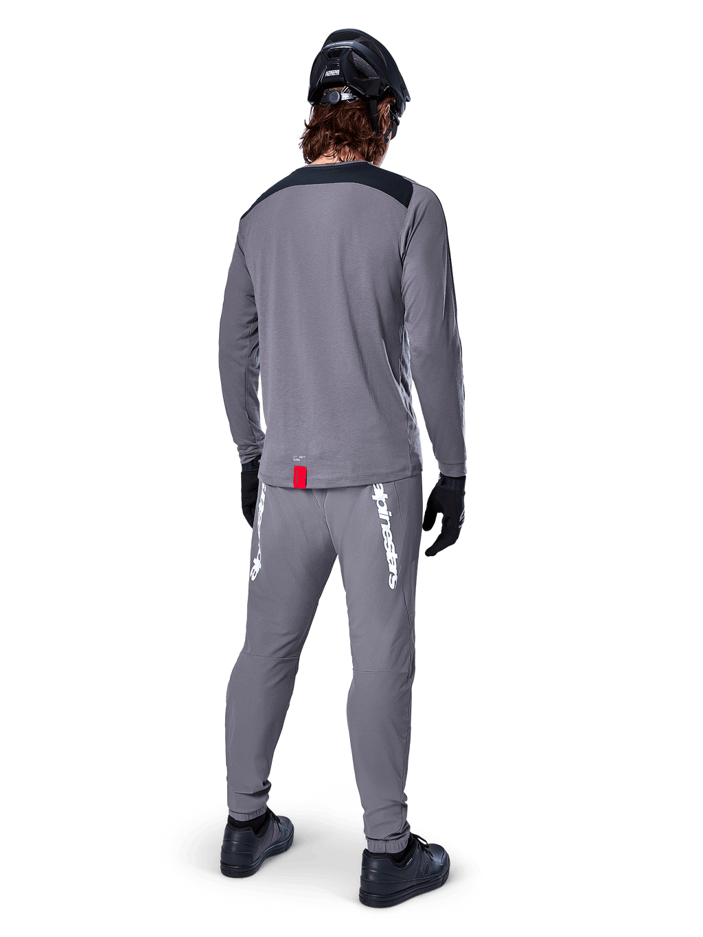 Jersey de manga larga Alpinestars A-Dura Dri Astar, gris oscuro, vista posterior del ciclista con pantalones a juego y casco negro, cuenta con paneles negros en los hombros y una pequeña etiqueta roja en el dobladillo, equipamiento técnico para ciclismo todoterreno