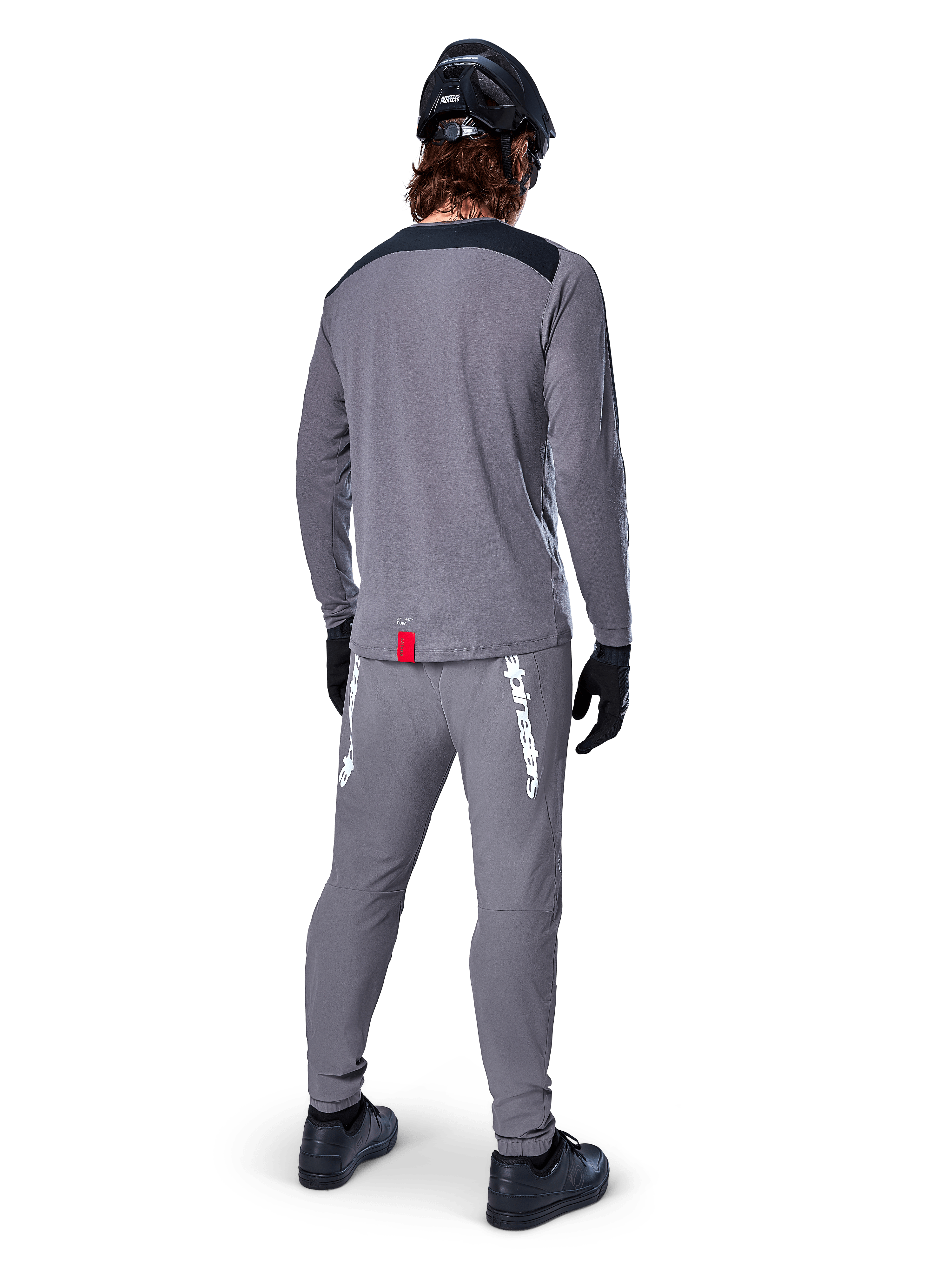 Jersey de manga larga Alpinestars A-Dura Dri Astar, gris oscuro, vista posterior del ciclista con pantalones a juego y casco negro, cuenta con paneles negros en los hombros y una pequeña etiqueta roja en el dobladillo, equipamiento técnico para ciclismo todoterreno