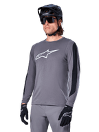 Jersey de manga larga Alpinestars A-Dura Dri Astar, gris oscuro con logotipo blanco en el pecho y paneles laterales de malla negra, jersey de ciclismo de montaña MTB, usado con casco negro, gafas de sol y pantalones grises a juego
