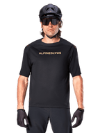 Ciclista de montaña con el jersey Alpinestars A-Aria Polartec Switch de manga corta en color negro, con logotipo dorado en el pecho, combinado con casco de MTB negro, gafas de sol oscuras de alto rendimiento y guantes a juego, equipamiento de ciclismo de montaña.