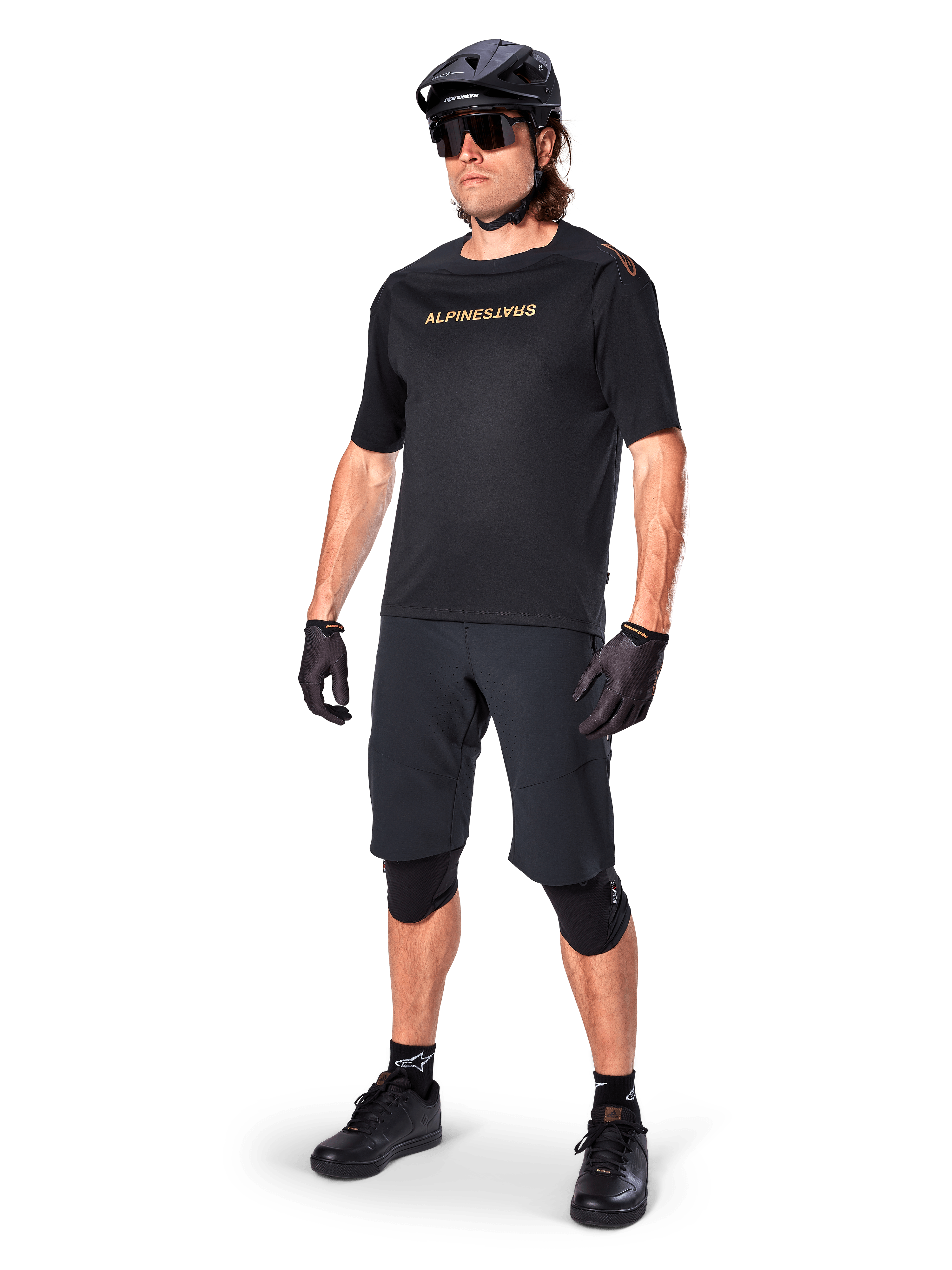Jersey de manga corta Alpinestars A-Aria Polartec Switch, negro, con logotipo en el pecho en dorado efecto espejo, lucido por un ciclista con pantalones cortos de ciclismo, casco y rodilleras negros, equipamiento técnico de MTB para rendimiento todoterreno