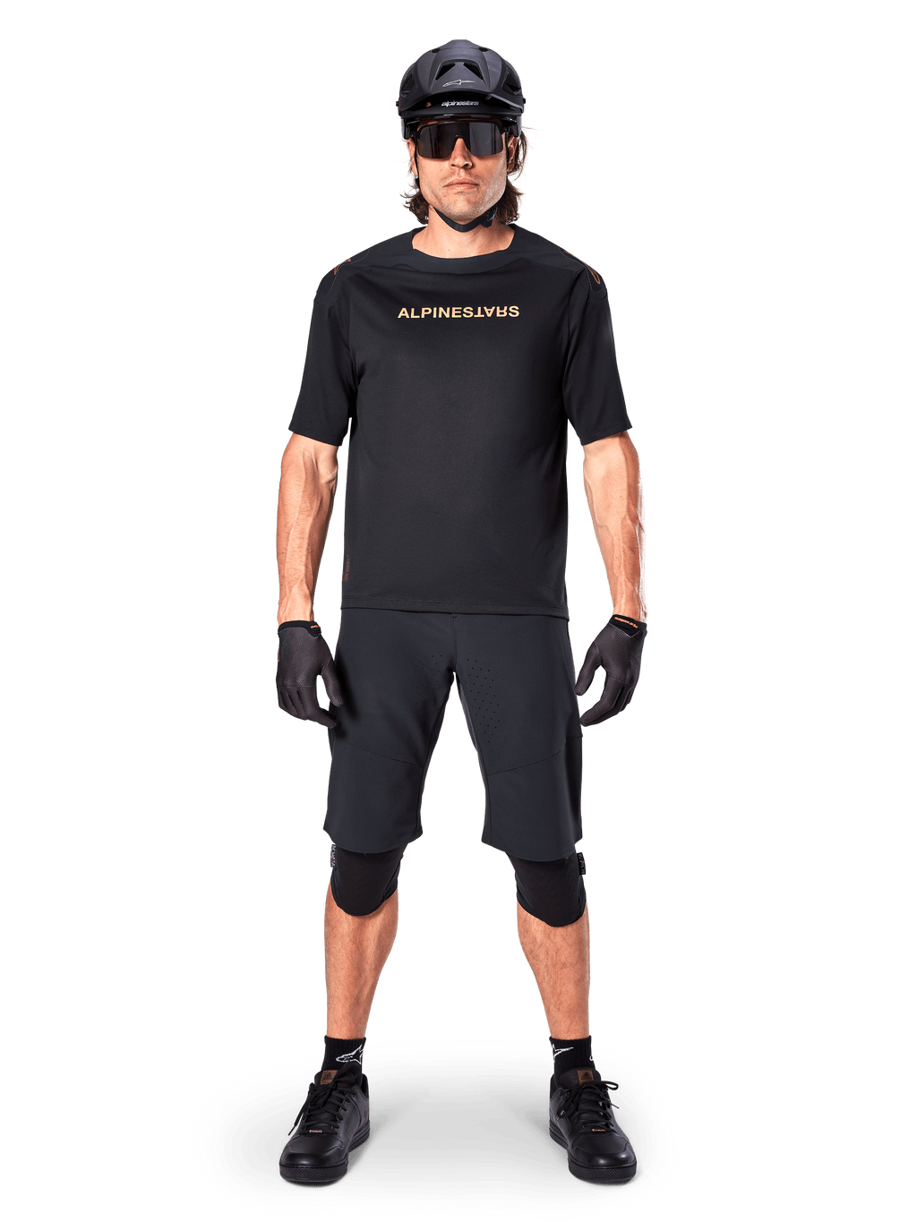 Alpinestars A-Aria Polartec Switch Jersey, jersey de MTB de manga corta, negro con letras en color canela, usado con equipamiento de mountain bike negro coordinado que incluye casco, pantalones cortos y rodilleras, ropa de ciclismo de alto rendimiento para off-road