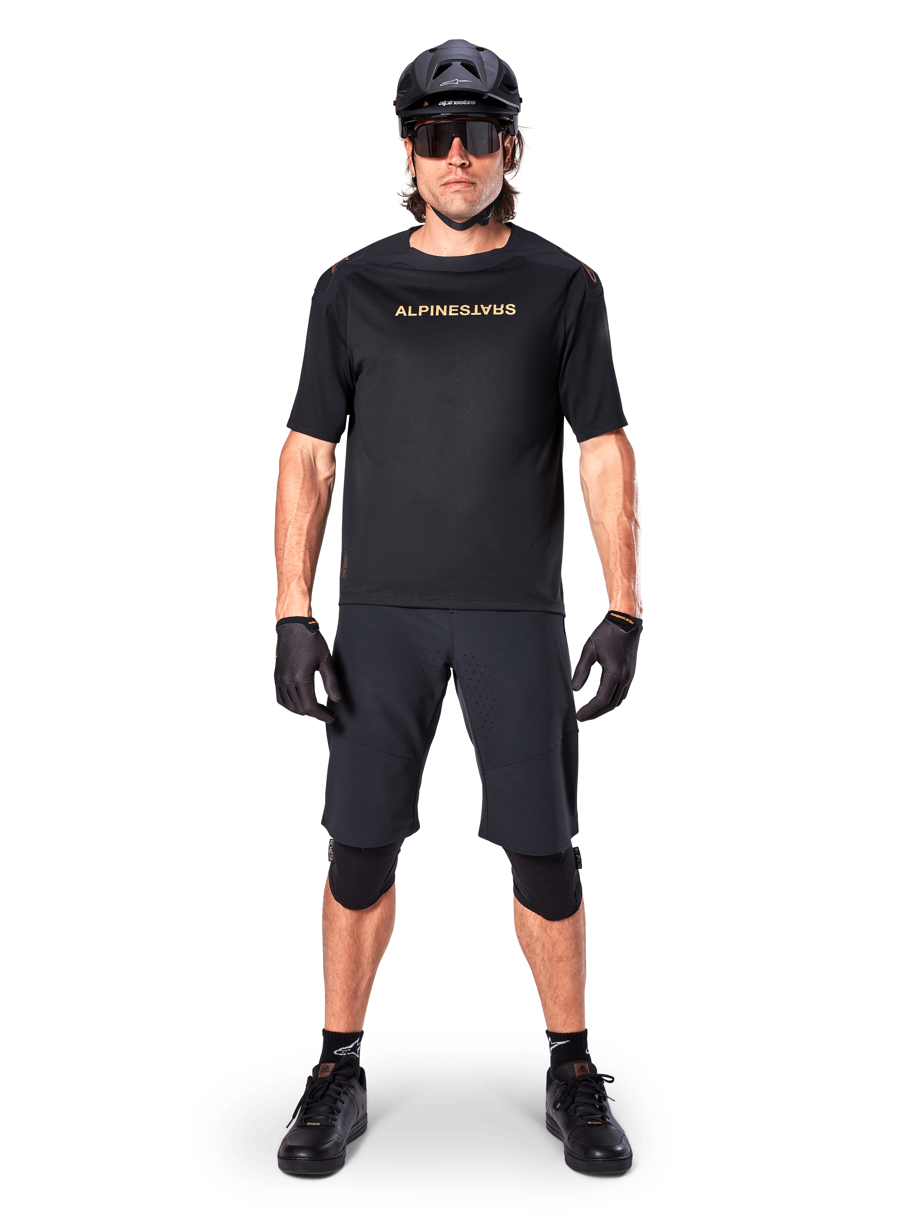 Alpinestars A-Aria Polartec Switch Jersey, jersey de MTB de manga corta, negro con letras en color canela, usado con equipamiento de mountain bike negro coordinado que incluye casco, pantalones cortos y rodilleras, ropa de ciclismo de alto rendimiento para off-road
