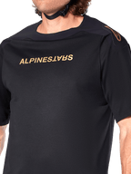 Alpinestars A-Aria Polartec Switch Jersey, camiseta de MTB de manga corta, negro, letras doradas de Alpinestars en el pecho y logotipo en el hombro, primer plano del torso superior que resalta el tejido técnico y el ajuste de alto rendimiento