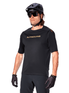 Alpinestars A-Aria Polartec Switch Jersey de manga corta, camiseta negra de ciclismo de montaña con la marca Alpinestars en dorado, usada con casco negro, gafas de sol y guantes, equipamiento técnico de MTB