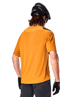 Jersey Alpinestars A-Aria Polartec Switch, camiseta de MTB de manga corta, color oro oscuro, vista posterior que muestra el ajuste de rendimiento, usado con casco negro y pantalones cortos grises, ropa de trail duradera y transpirable