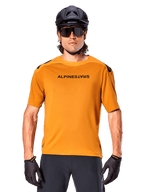 Jersey Alpinestars A-Aria Polartec Switch de manga corta, color oro oscuro, ciclista de montaña con casco negro y gafas de sol, ropa técnica de alto rendimiento para senderos de MTB