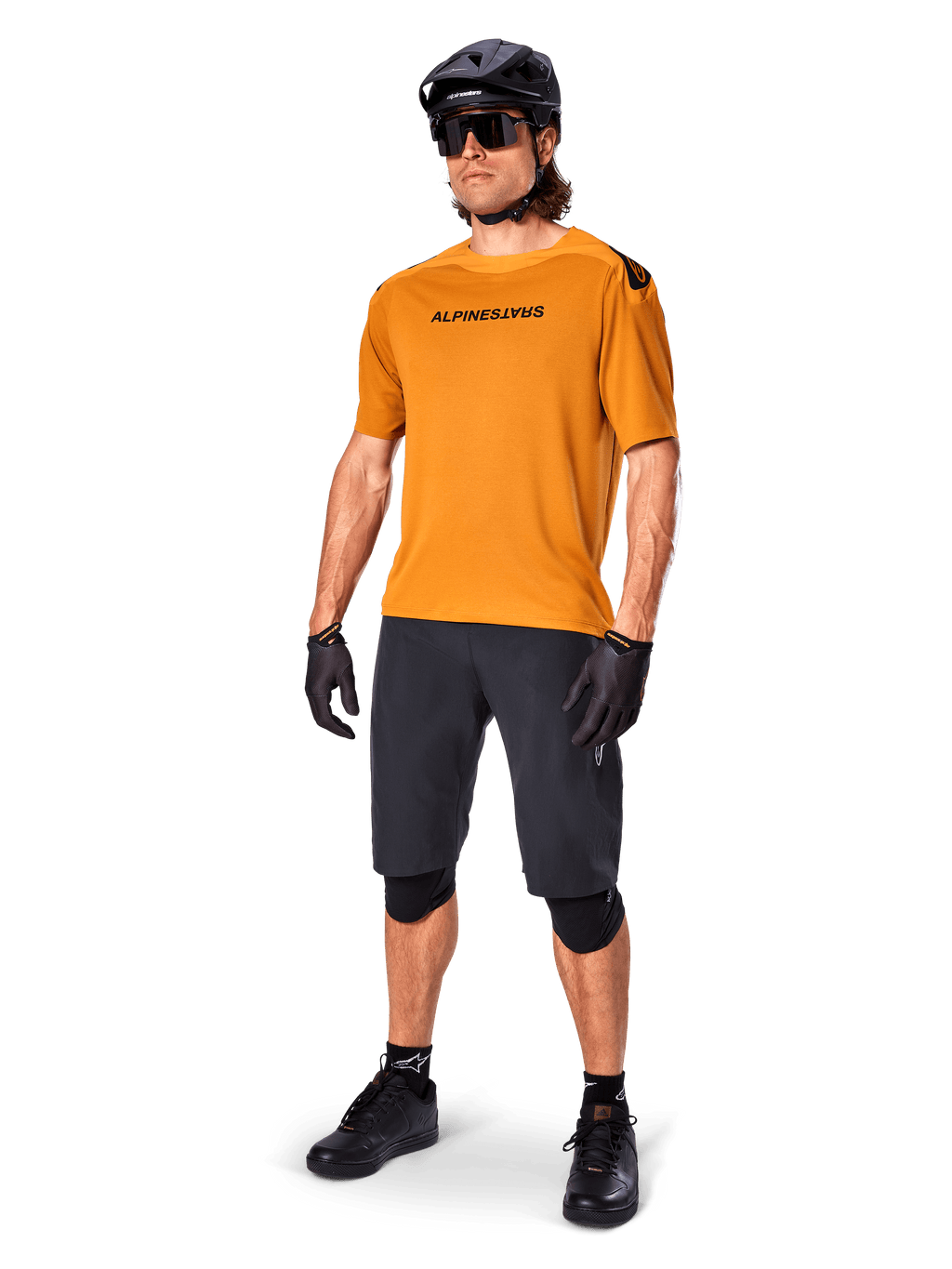 Ciclista masculino con el jersey Alpinestars A-Aria Polartec Switch en color Dark Gold, jersey de MTB de manga corta, combinado con pantalones cortos de ciclismo negros, rodilleras, casco negro y guantes, equipamiento profesional de bicicleta de montaña para trail