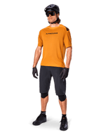 Ciclista masculino con el jersey Alpinestars A-Aria Polartec Switch en color Dark Gold, jersey de MTB de manga corta, combinado con pantalones cortos de ciclismo negros, rodilleras, casco negro y guantes, equipamiento profesional de bicicleta de montaña para trail