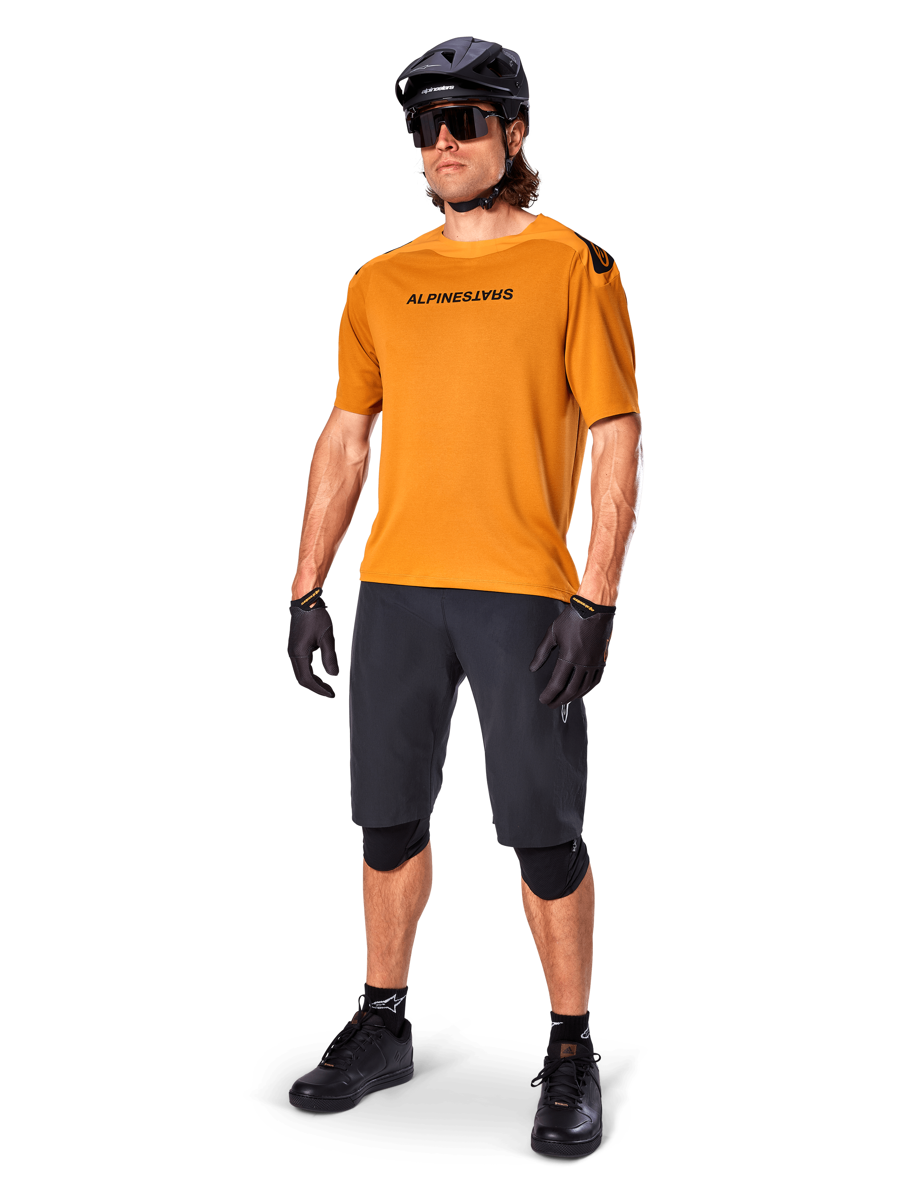 Ciclista masculino con el jersey Alpinestars A-Aria Polartec Switch en color Dark Gold, jersey de MTB de manga corta, combinado con pantalones cortos de ciclismo negros, rodilleras, casco negro y guantes, equipamiento profesional de bicicleta de montaña para trail
